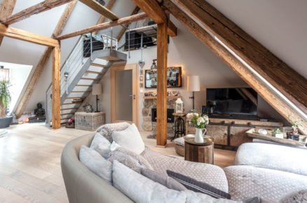Obývací pokoj ve venkovském stylu. Do patra vede kovové schodiště, pokoj je zařízený masivním dřevěným nábytkem, součástí pokoje jsou dřevěné trámy