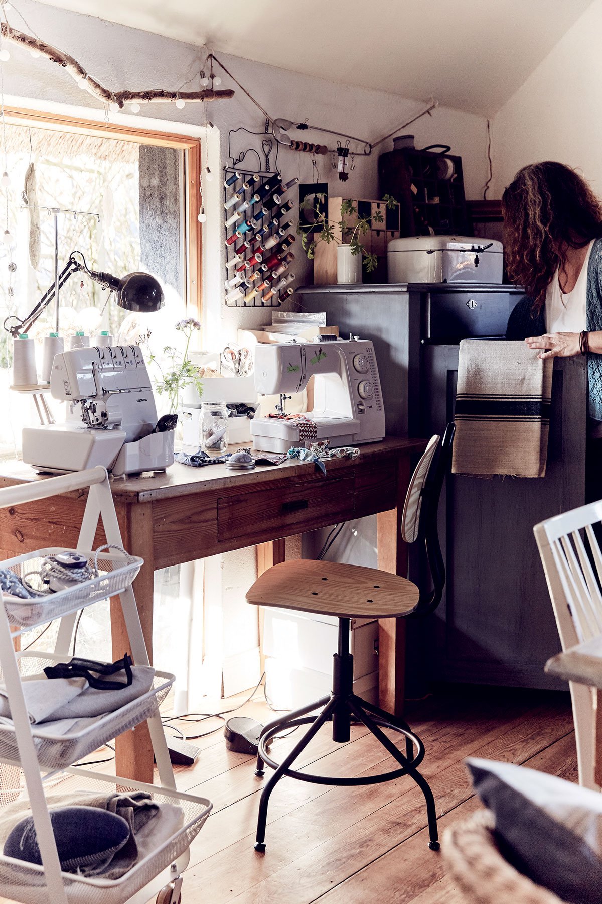 pracovní stůl ve venkovském stylu se šicím strojem, organizérem na kolečkách a otáčavou dřevěnou industriální židlí