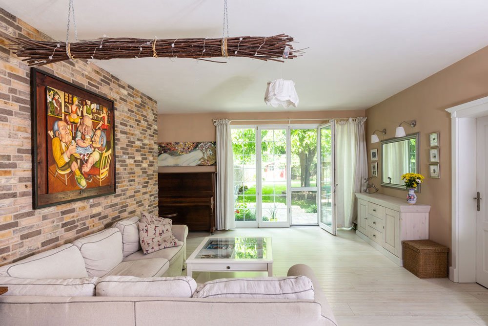 obývací pokoj se vstupem do zahrady ve venkovském stylu s bílou pohovkou, skleněným stolkem, bílou komodou, klavírem a cihlovou zdí