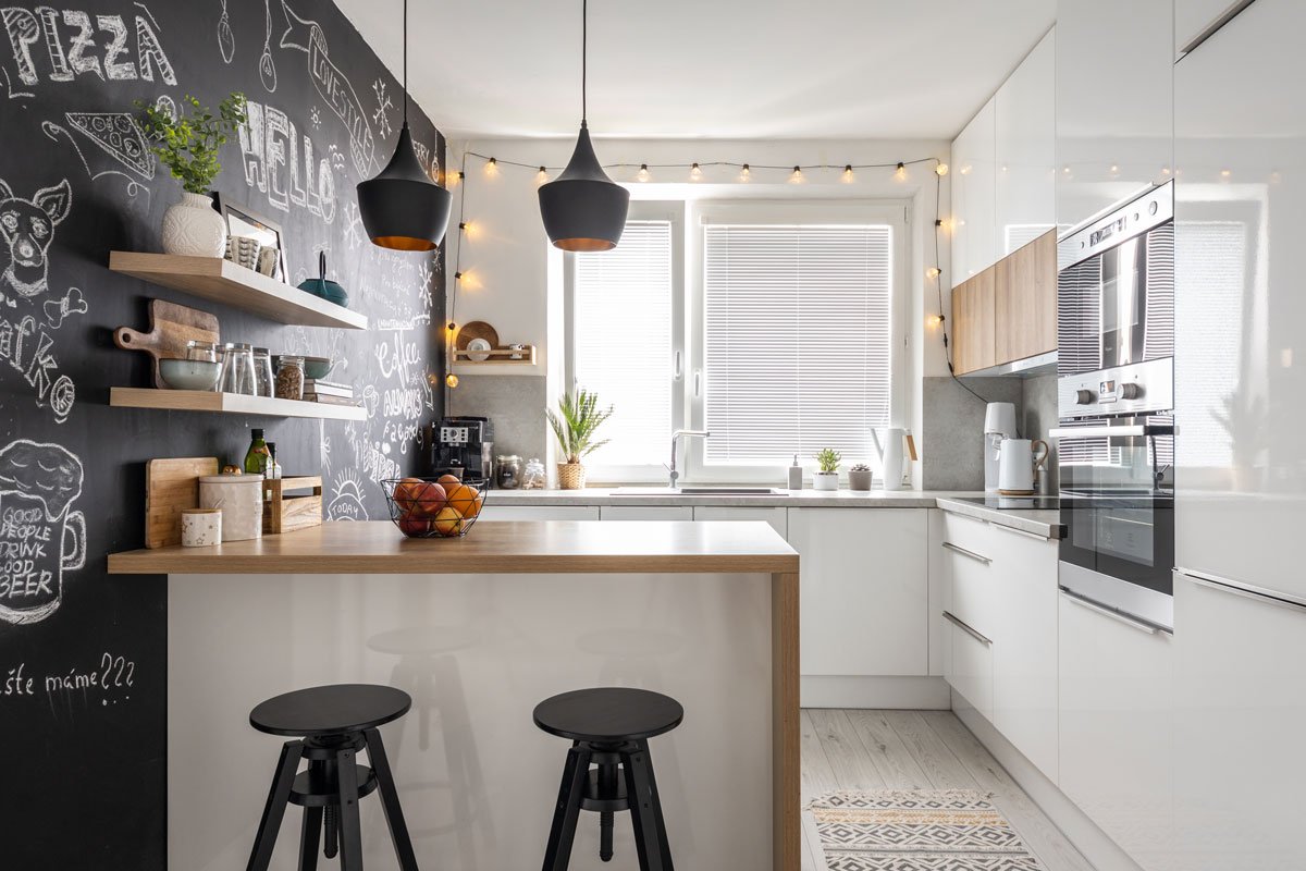 Kuchyně s bílou kuchyňskou linkou, ostrůvkem, barovými židlemi a černou tabulovou stěnou