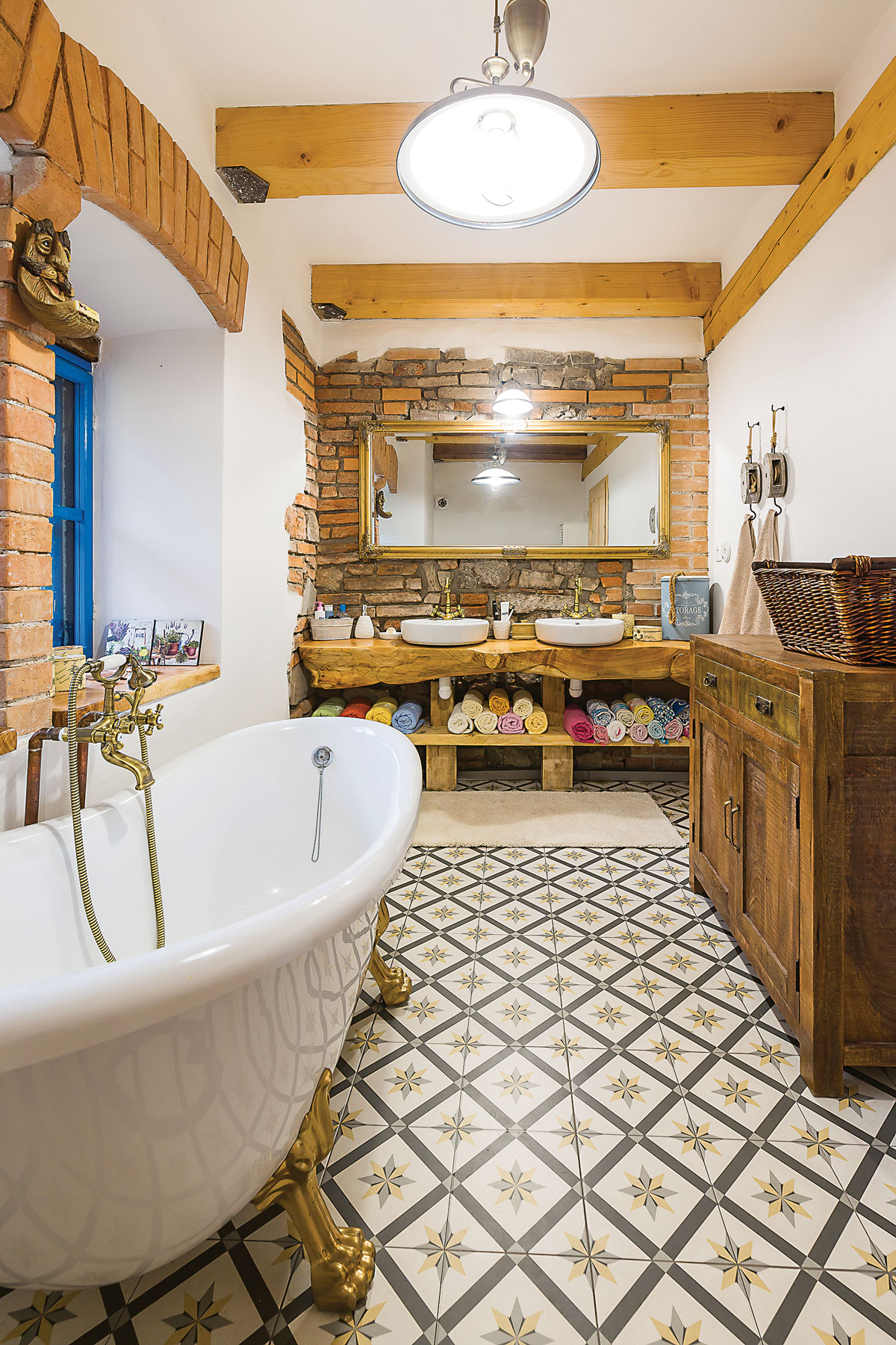 koupelna ve venkovském stylu s vanou na zlatých nožičkách, přiznanou cihlovou zdí a umývadly položenými na masivním kusu opracovaného dřeva