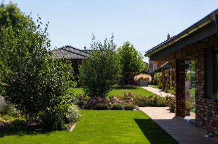 Pohled na rodinný dům, zahradní kuchyň, trávnik s okrasnými záhony a ihřiště pro děti