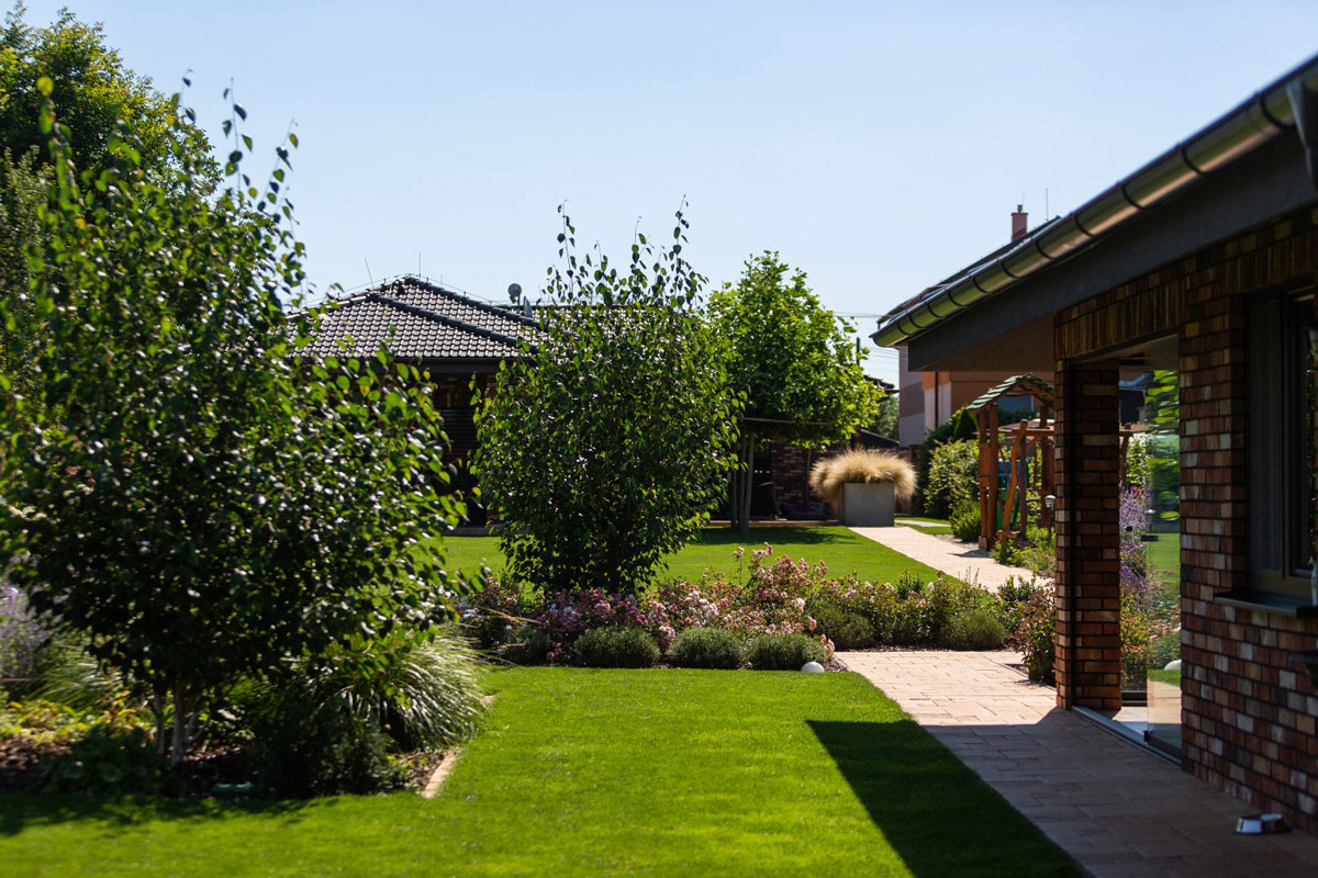 Pohled na rodinný dům, zahradní kuchyň, trávnik s okrasnými záhony a ihřiště pro děti