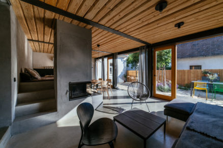moderní přístavba ke chalupě s obývacím pokojem s industriálním nádychem, na zdech je betonová omítka, strop tvoří dřevěné trámy a prosklená stěna vede na terasu