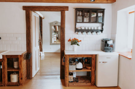Venkovský interiér kuchyně s otevřenými dřevěnými policemi, masivní dřevěnou zárubní a trámy na stropě