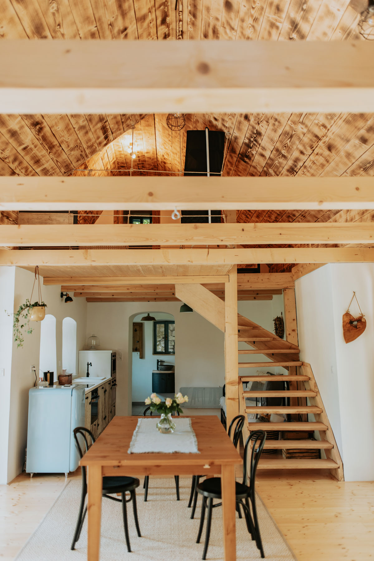 venkovský interiér s dřevěným stolem a černými židlemi, v pozadí je kuchyň, odkud vede dřevěné schodiště