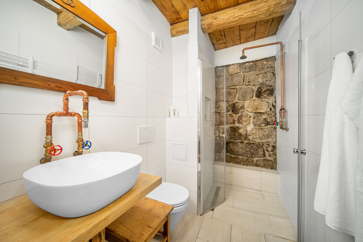 koupelna v industriálním stylu na chalupě, ve sprchovém koutě je přiznána kamenná zeď a zajímavým prvkem jsou industriální baterie