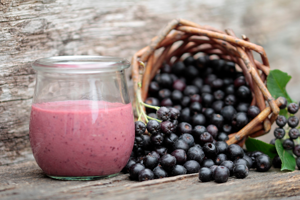 Sklenička hustého růžovo-fialového smoothie z aronie před proutěným košíkem, ze kterého se sypou čerstvé černé bobule; rustikální dřevěné pozadí.