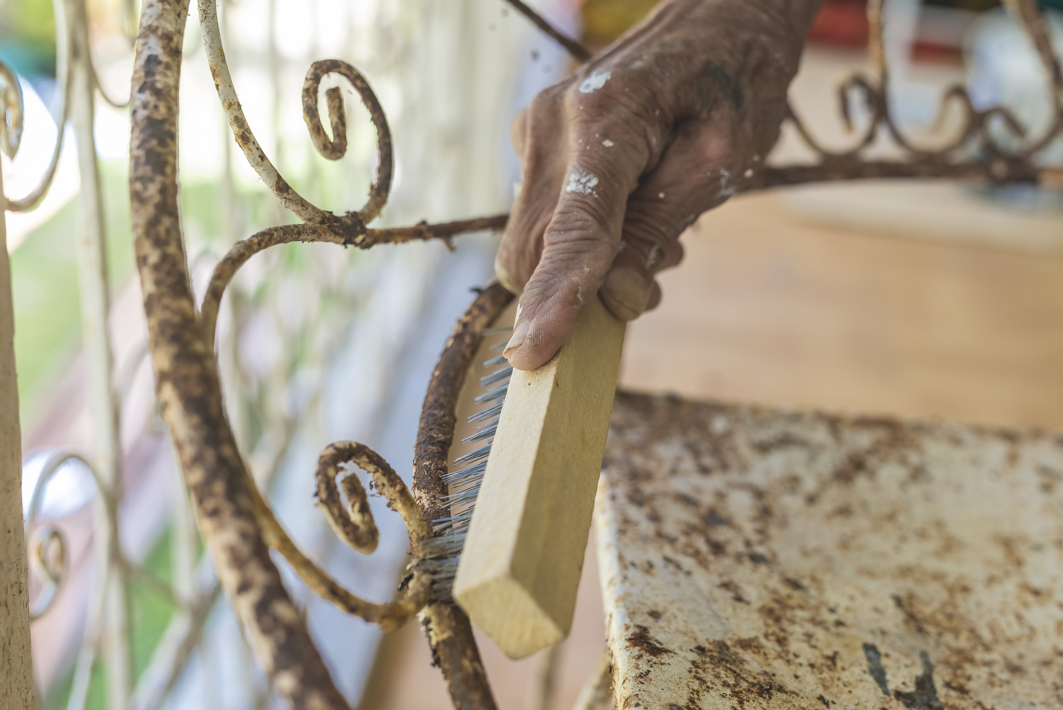 A man uses a steel brush to scrape off old paint and rust from an old wrought iron chair. Furniture repair and restoration concept.