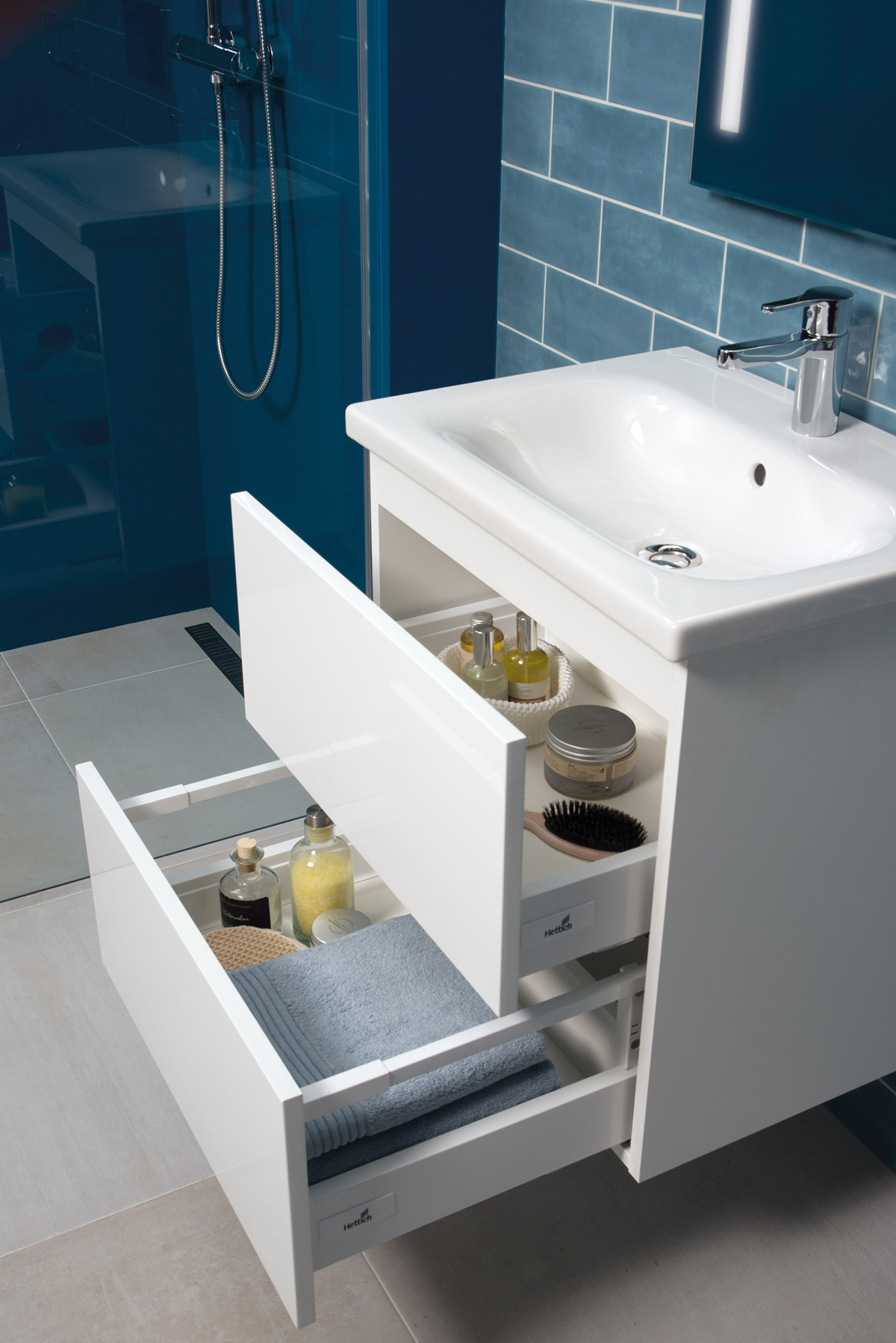 Mio N Jika (18) Otevřená zásuvka bílé koupelnové skříňky s ručníky a kosmetikou.