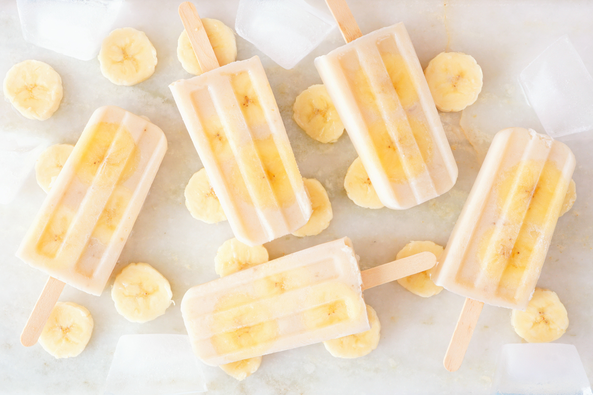 bananove nanuky Banánové domácí nanuky s kousky banánu, naaranžované s plátky banánu a kostkami ledu na bílém povrchu.