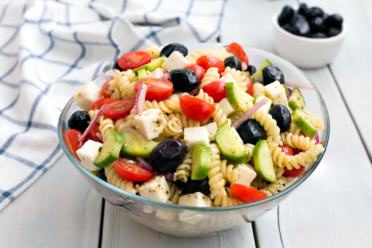 recky salat Skleněná miska s těstovinovým salátem, cherry rajčaty, okurkou, olivami, fetou a červenou cibulí na světlém podkladu.