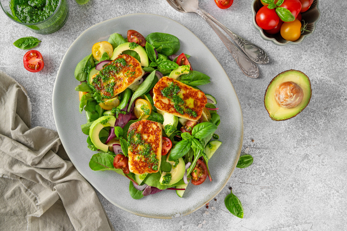 halloumi salat Talíř salátu s grilovanými plátky halloumi, plátky avokáda, cherry rajčaty, červenou cibulí a zelenými listy, ozdobený pestem.