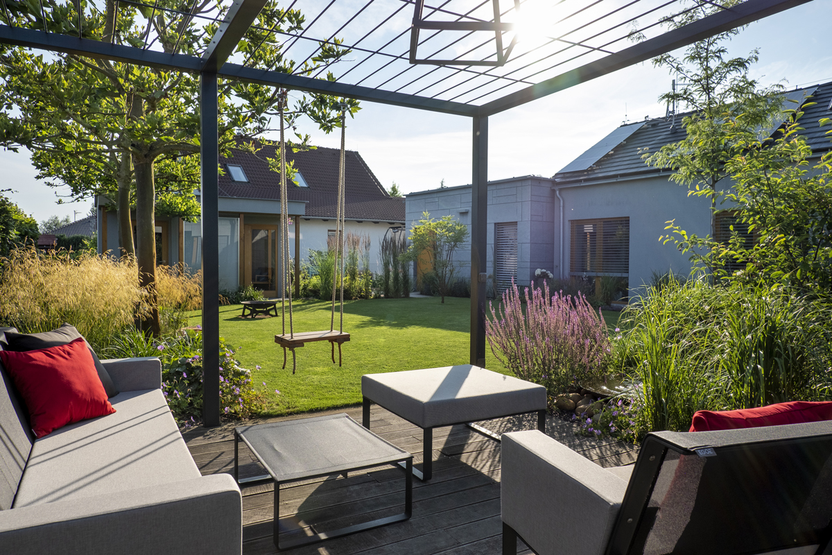pergola Pohled z terasy s pergolou do rozkvetlé zahrady s trávníkem, houpačkou a zahradním nábytkem, slunce vytváří dlouhé stíny.