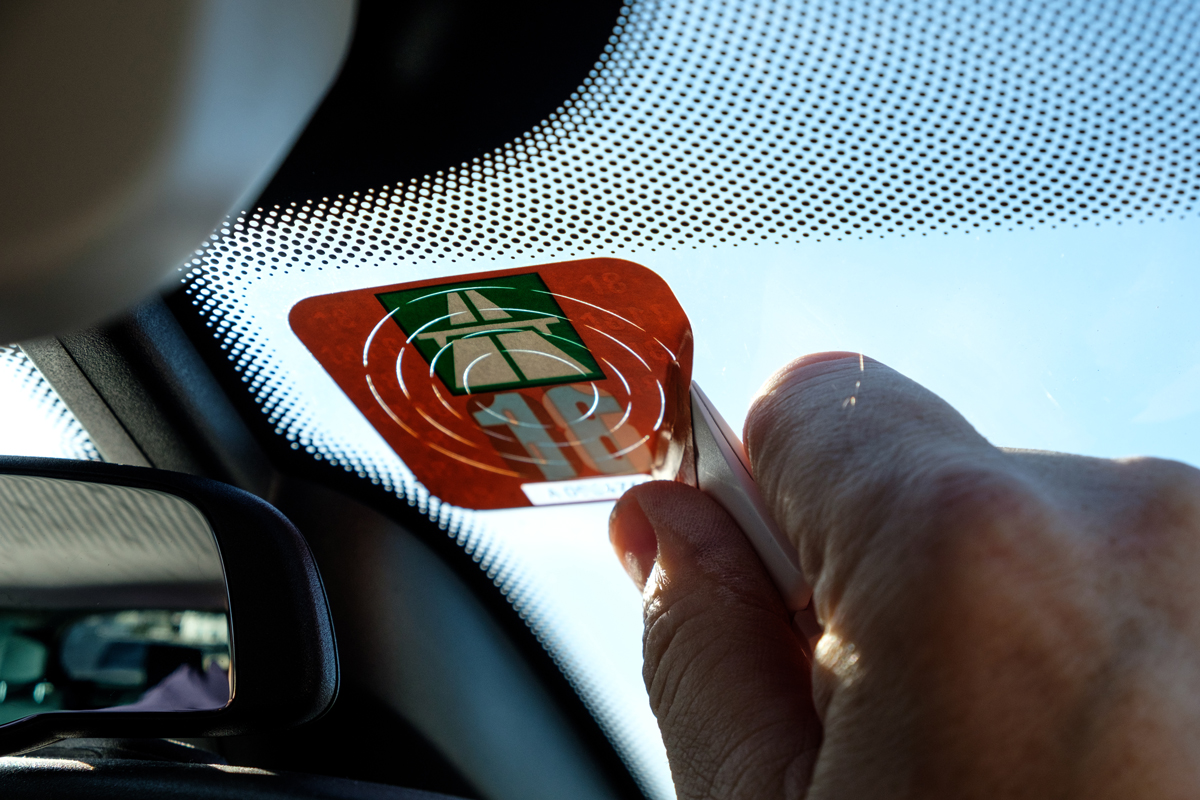 Ruka přilepuje dálniční známku na čelní sklo automobilu s výhledem na modrou oblohu.