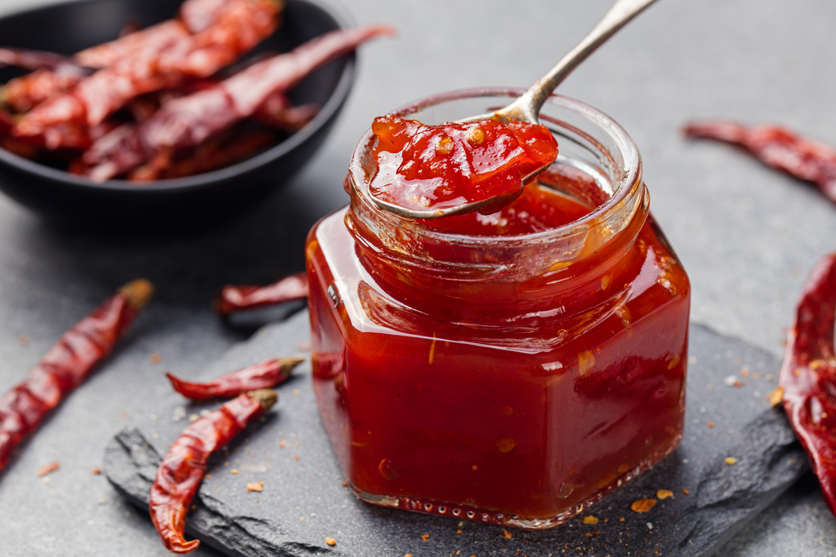 Sklenice s domácím rajčatovým čatní, lžička s čatní v popředí, v okolí sušené červené chilli papričky.