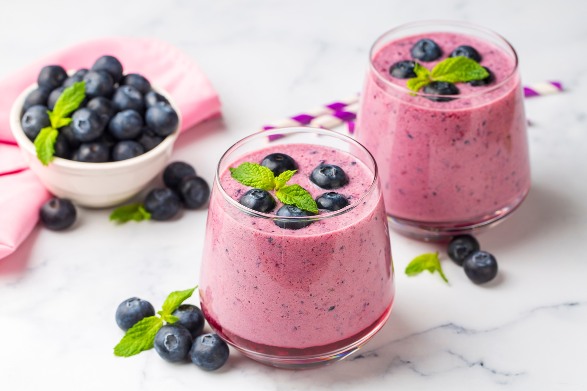 Dvě skleničky hustého borůvkového smoothie z borůvek, banánu a jogurtu, ozdobené čerstvými borůvkami a mátou; v pozadí miska borůvek. Jemně sladké, lehce nakyslé a sametové, ideální pro rychlý ranní boost.