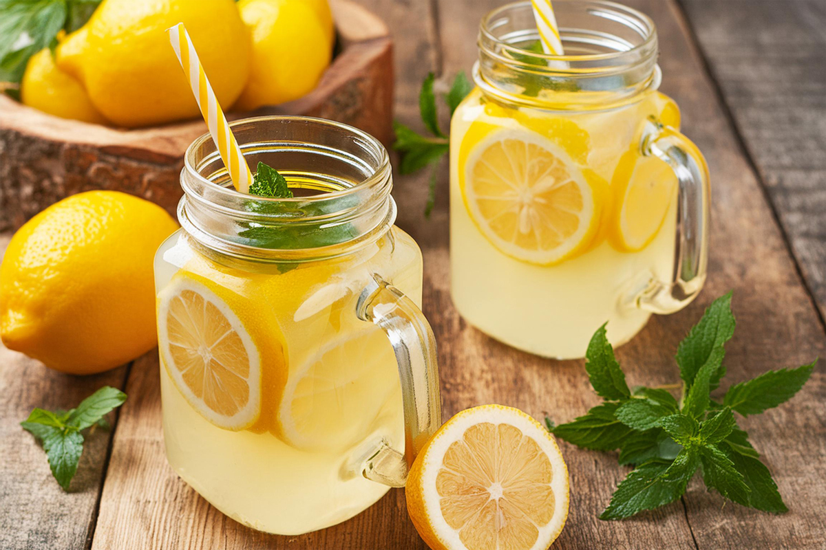 Dvě zavařovací sklenice s domácí citronovou limonádou, plátky citronu, mátou a brčky na dřevěném stole; okolo celé citrony a snítky máty.