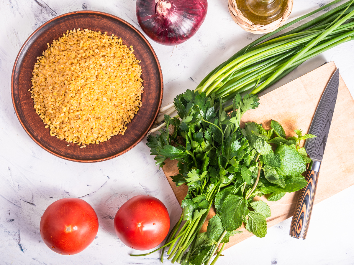 ingredience Ingredience na vaření tabbouleh - bulgur v dřevěné misce, rajčata, jarní cibulka a petrželka.