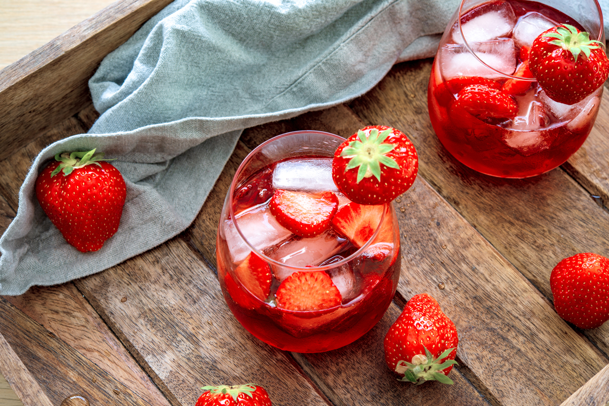 Sklenice jahodové limonády s ledem a plátky jahod na dřevěném tácu, kolem čerstvé jahody a šedý lněný ubrousek.