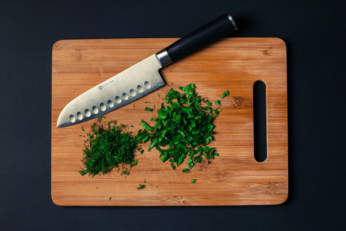 Santoku nůž s grantony krájí čerstvé bylinky na dřevěném prkénku.