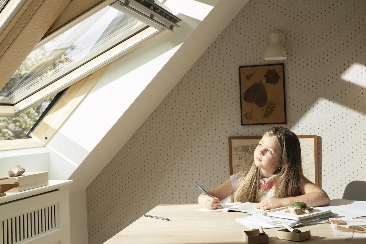 Podkrovní pokoj s dívkou u psacího stolu, střešní okno VELUX vpouští maximum denního světla, ideální prostředí pro učení a kreativitu.