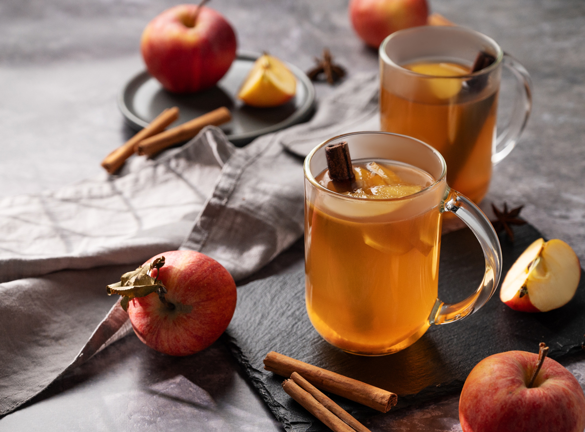 Dvě sklenice jablečného cideru s tyčinkou skořice a čerstvými jablky kolem.