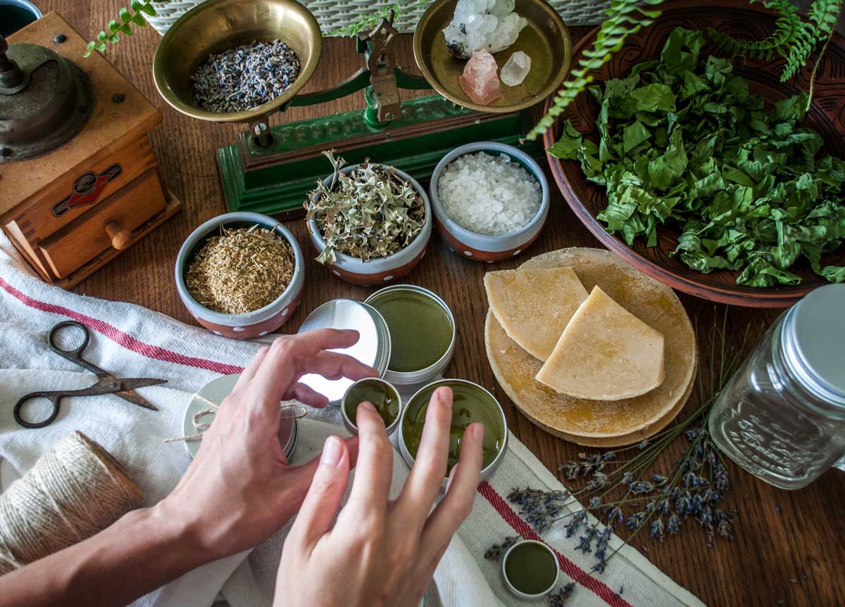 Výroba bylinné masti proti kašli s použitím sušených bylin, včelího vosku a přírodních olejů na stole plném léčivých ingrediencí.