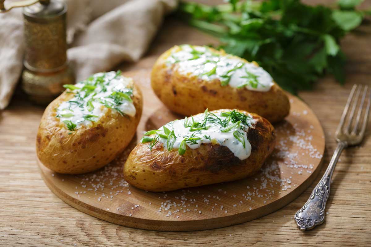 Pečené brambory s bylinkami a smetanou naservírované na dřevěném prkénku.