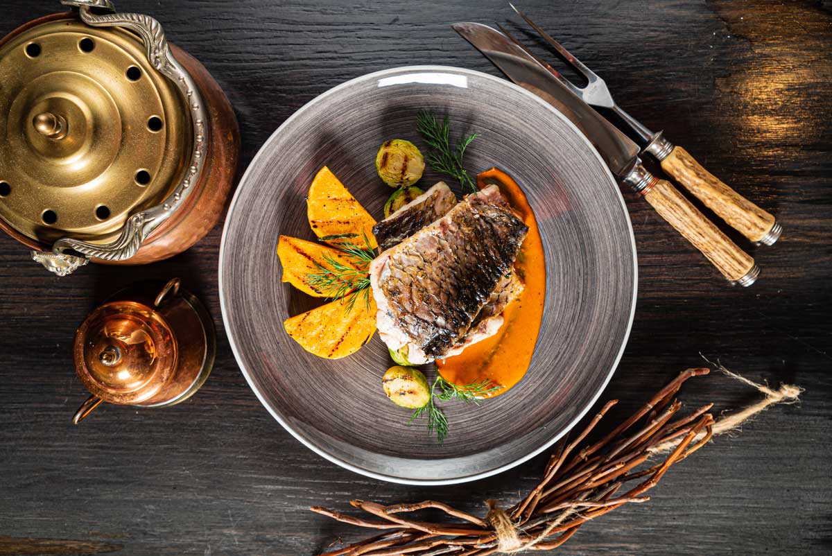 Delikátní porce kapra na jemné omáčce s grilovanou zeleninou působí jako moderní pocta tradiční kuchyni.