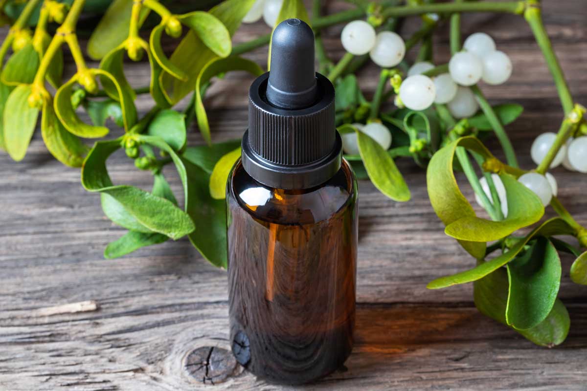 Léčivá tinktura z jmelí v hnědé lahvičce položená na dřevěném stole se snítkami jmelí v pozadí.