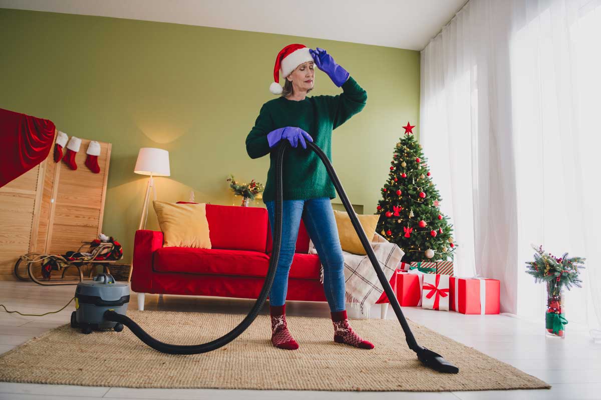 Žena v santa čepici uklízí obývák před Vánoci a vypadá unaveně z předvánočního shonu.