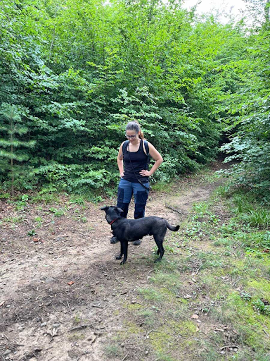 Žena stojí v lese se psem, který ji sleduje a reaguje na její podněty během společné procházky.