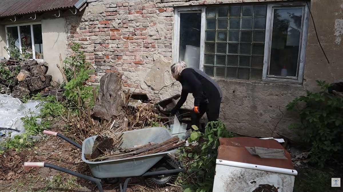 Žena třídí dřevěný a kovový odpad u staré zdi domu a připravuje jej k odvozu.