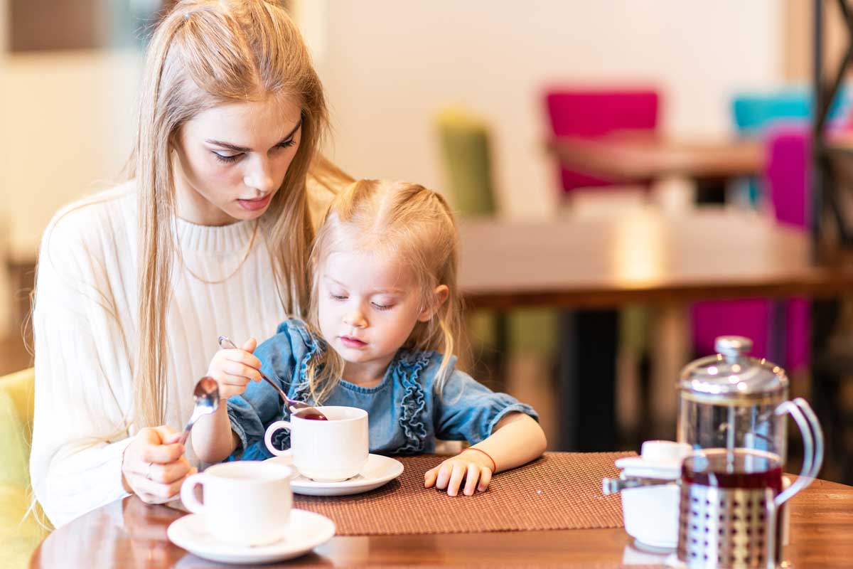 Malé dítě sedí u stolu s hrnkem a dospělá osoba ho doprovází