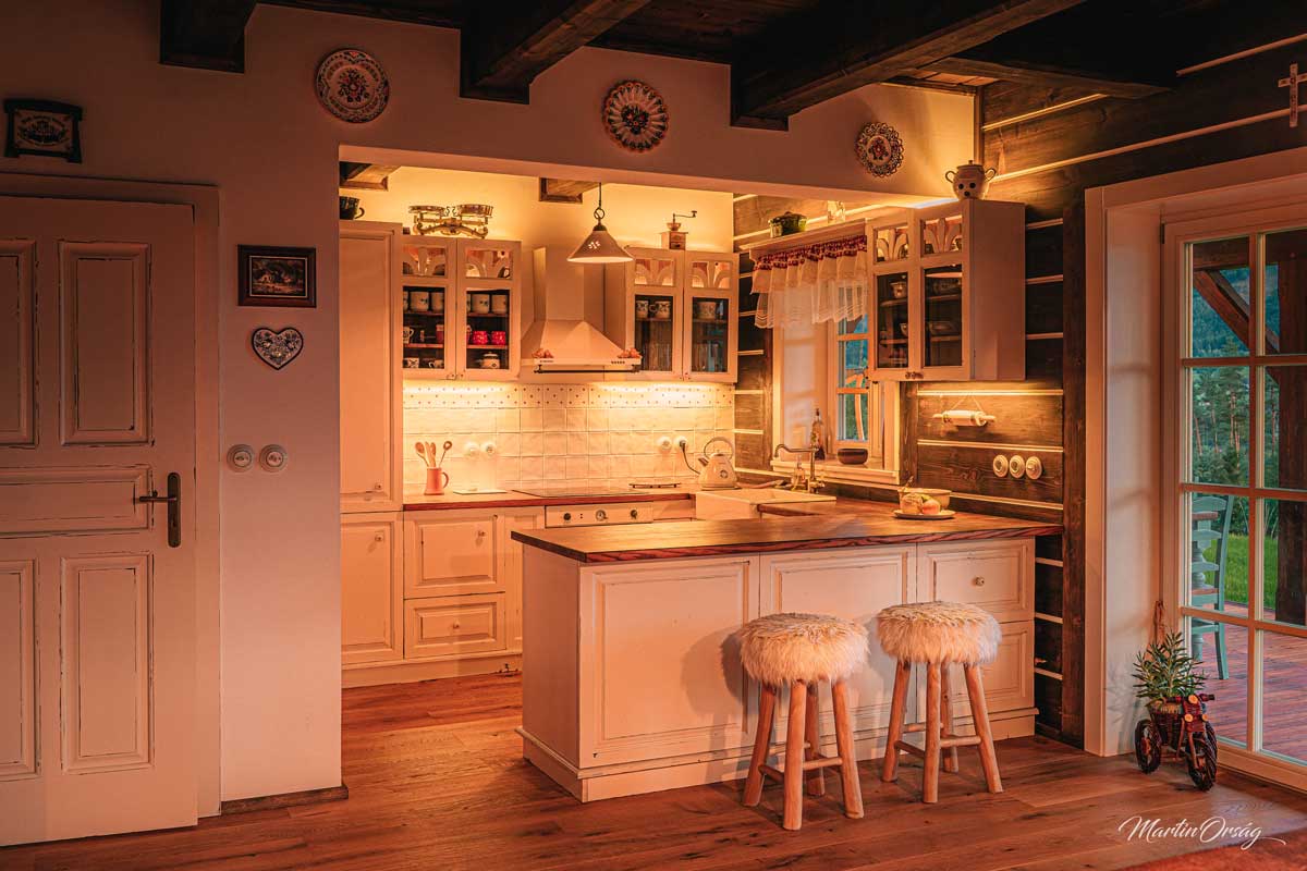 Bílá kuchyně v roubence s dřevěnými prvky a jemným podsvícením vytvářejícím atmosféru.