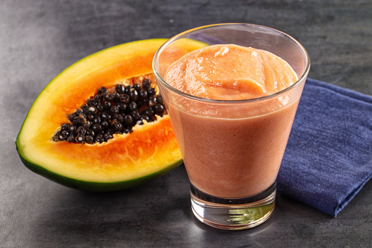 Sklenice hustého ovocného smoothie stojí vedle rozpůlené papáji s tmavými semínky.
