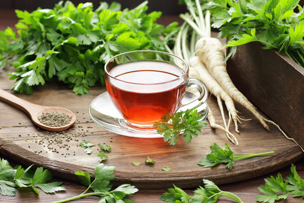 Šálek petrželového čaje je položený na dřevěném podnose obklopený čerstvou petrželí a kořenovou zeleninou.