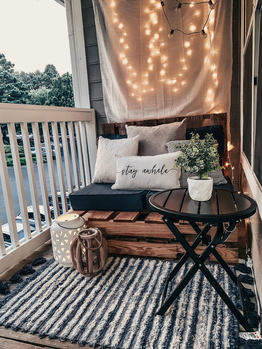 Malý balkon s posezením, dekoračními světýlky, kobercem a polštáři připravený k večernímu odpočinku.