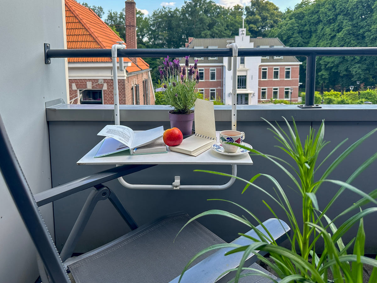 Balkon s přenosným stolkem, kávou, knihou a květinou využitý jako klidné místo k rannímu posezení.