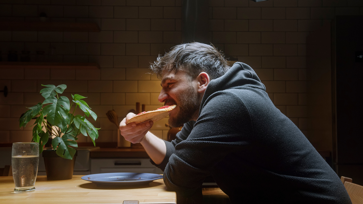 Muž jí v noci pizzu u kuchyňského stolu, což ilustruje, jak večer a v noci roste chuť na těžká a nezdravá jídla.