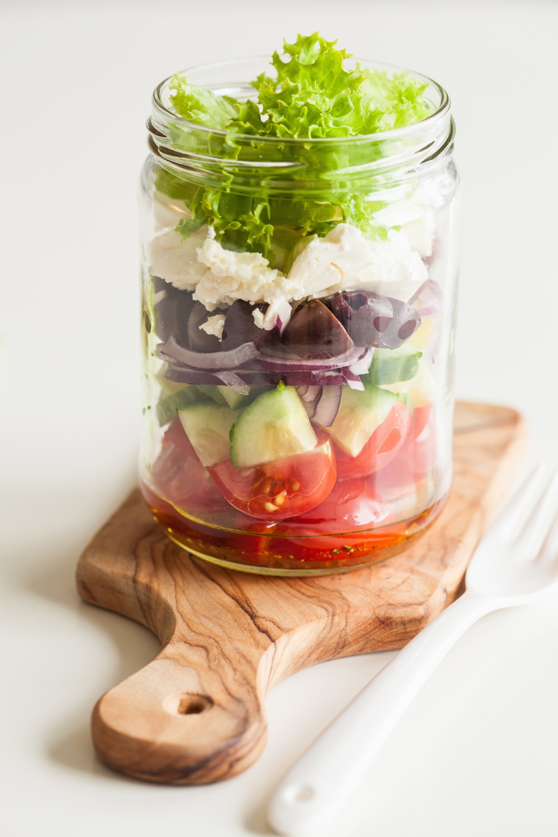 recky salat do sklenky Řecký salát ve sklenici s vrstvami dresinku, rajčat, okurky, oliv, červené cibule, sýra feta a křupavého salátu připravený jako praktický oběd do práce.