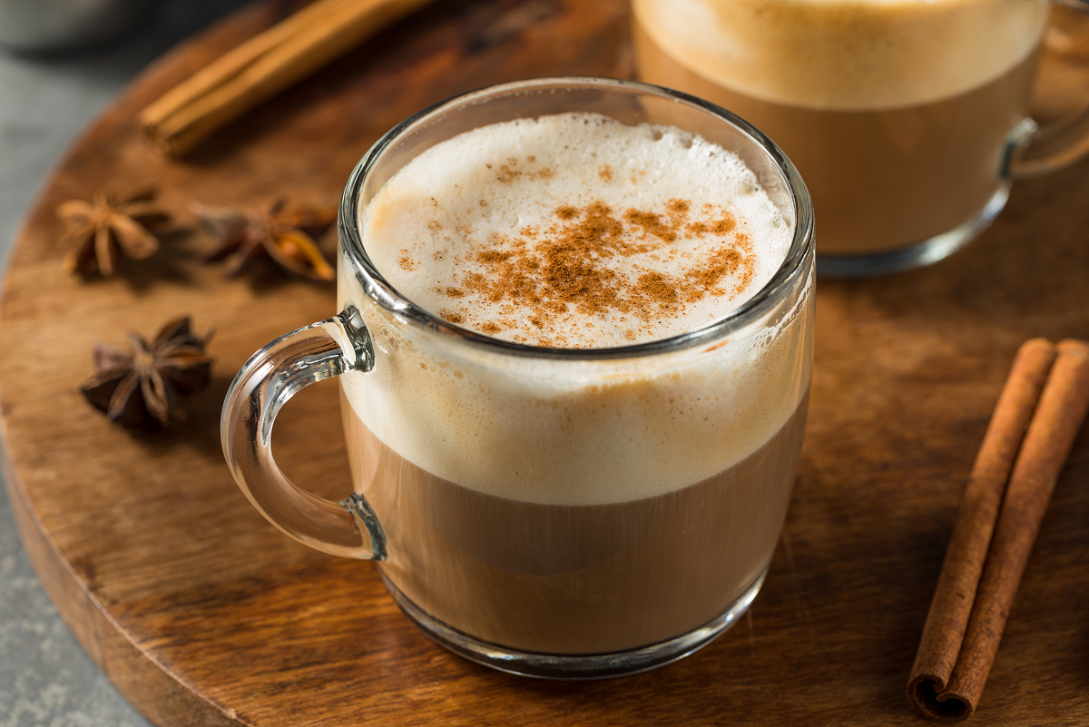 Chai latte s bohatou mléčnou pěnou posypanou skořicí ve skleněném hrnku, kolem kterého leží svitky skořice a badyán.