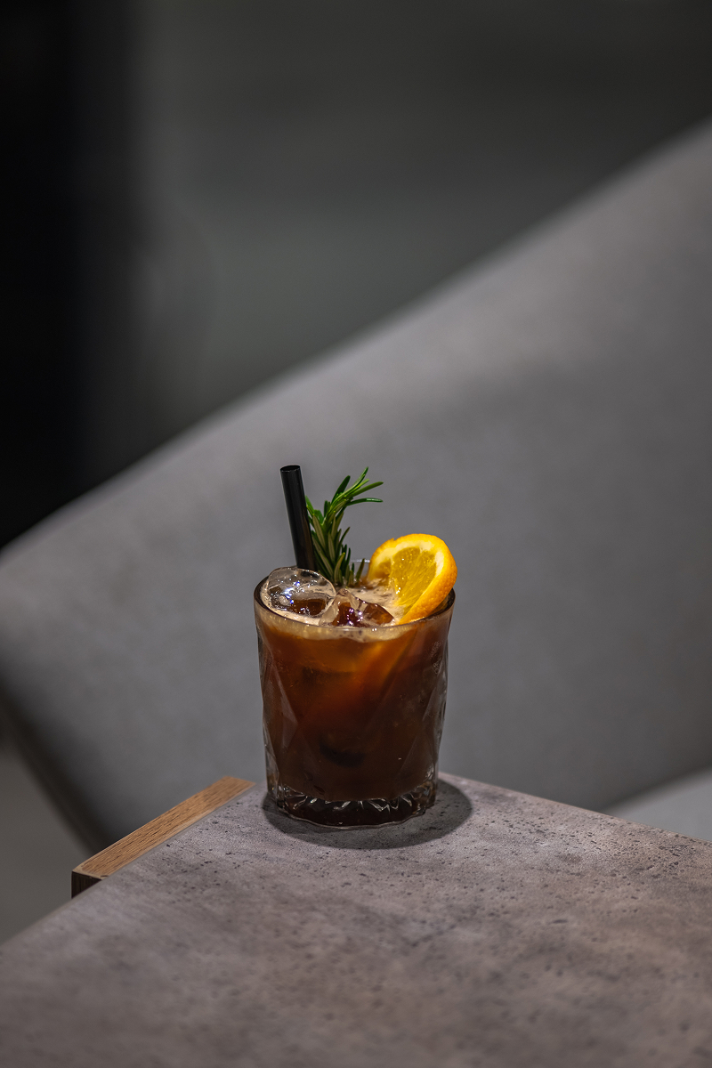Sklenice tmavé ledové kávy cold brew s kostkami ledu, plátkem pomeranče a snítkou rozmarýnu na šedém stolku.
