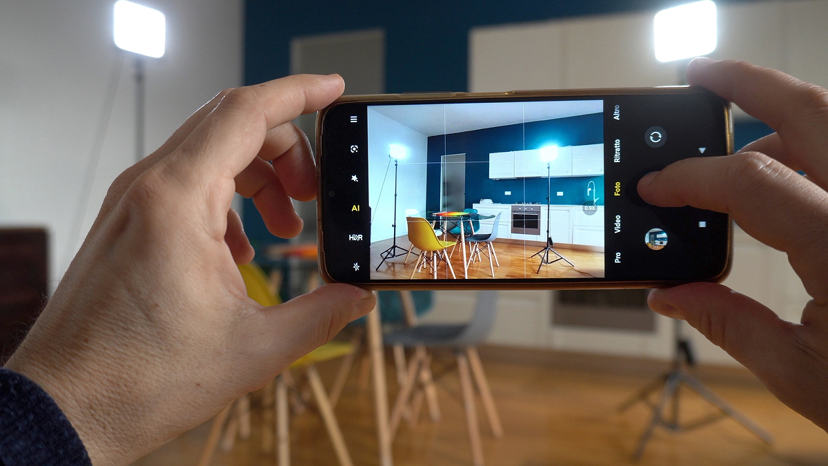 fotka pro AI 1 Fotografování moderní kuchyně mobilním telefonem, na jehož displeji je vidět bílá kuchyňská linka, jídelní stůl a tmavě modrá stěna.
