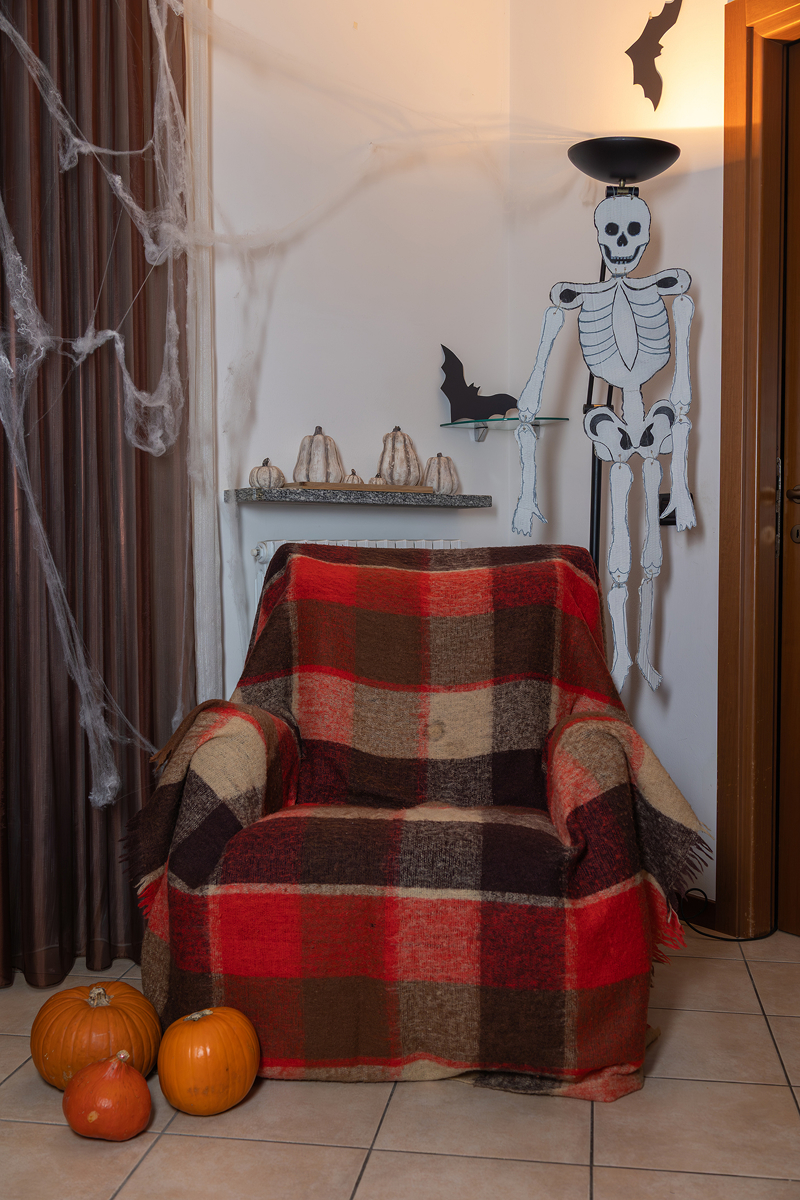 halloween kreslo 1 Křeslo s červenou kostkovanou dekou v halloweensky vyzdobeném koutku s papírovým kostlivcem, pavučinami, netopýry a dýněmi.
