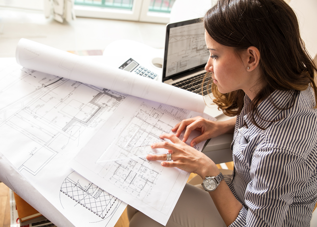 interierovy designer 1 Interiérová designérka u pracovního stolu prohlíží rozměrné architektonické výkresy a půdorysy s notebookem v pozadí.