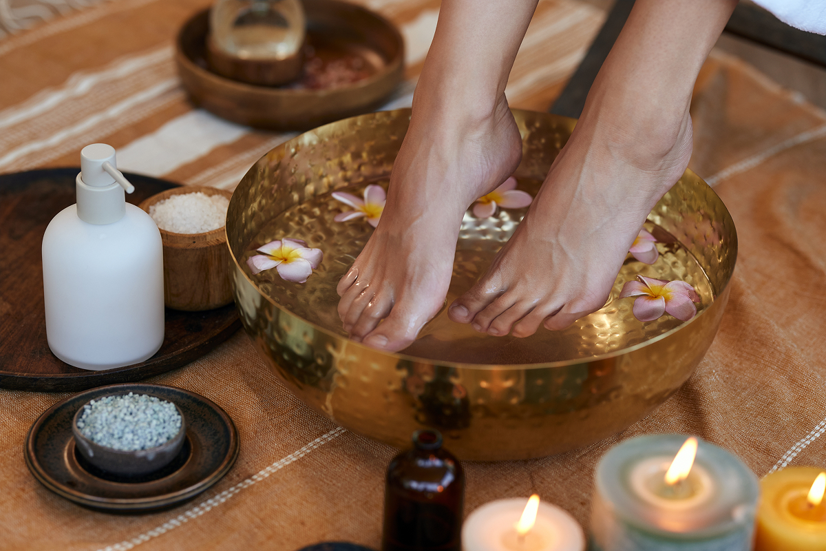 koupel nohou 1 Žena si namáčí chodidla ve zlaté kovové míse s vodou a růžovými květy, kolem které jsou rozmístěné koupelové soli, kosmetika a zapálené svíčky.