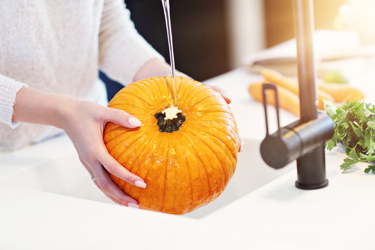 myti dyne 1 Oplachování celé oranžové dýně pod tekoucí vodou v kuchyňském dřezu před vyřezáváním halloweenské dekorace.