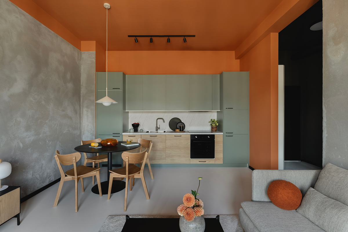 oranzovy strop 1 Moderní obývací pokoj propojený s kuchyní, kde výrazný oranžový strop doplňuje oranžové stěny, zelenou kuchyňskou linku a světlý nábytek.